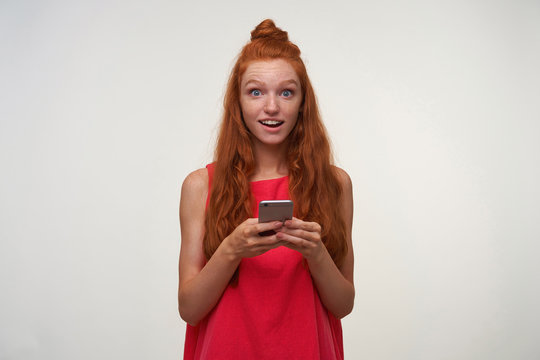 Surprised Young Lovely Female Wearing Her Red Hair In Knot, Standing Over White Background Without Makeup, Holding Smartphone In Hands And Looking To Camera With Raised Eyebrows And Round Eyes