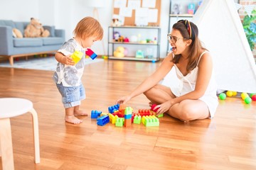 Beautiful teacher and toddler playing with building blocks around lots of toys at kindergarten