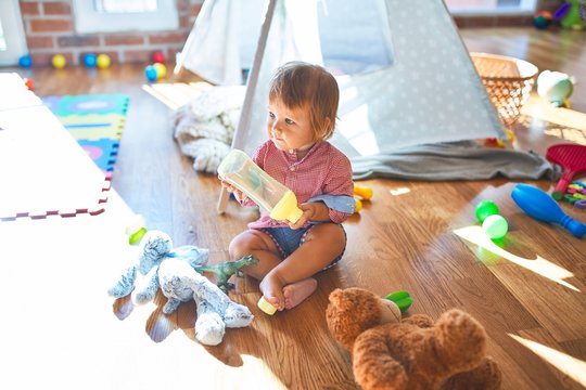 Adorable toddler holding feeding bottle around lots of toys at kindergarten