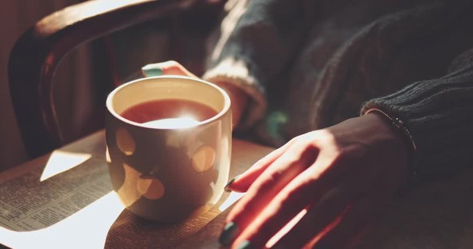 Woman Holding Steaming Cup of Tea or Coffee and Reading a Book, Enjoying Cozy Morning. SLOW MOTION, CLOSE UP. Unrecognizable Girl hands with a mug of hot drink indoors. Cinematic light. Lens Flare.