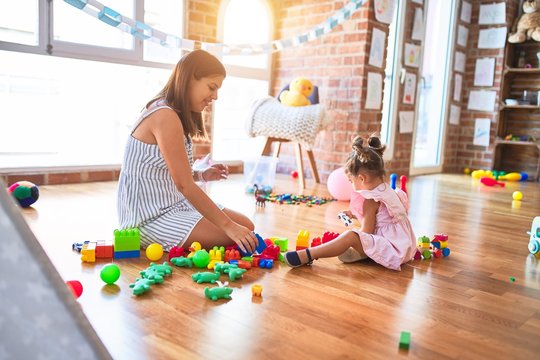 Young beautiful teacher and toddler sitting on the floor playing with building blocks toy at kindergarten