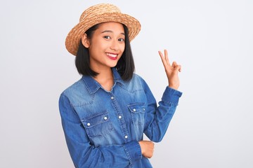 Young beautiful chinese woman wearing denim shirt and hat over isolated white background smiling with happy face winking at the camera doing victory sign. Number two.