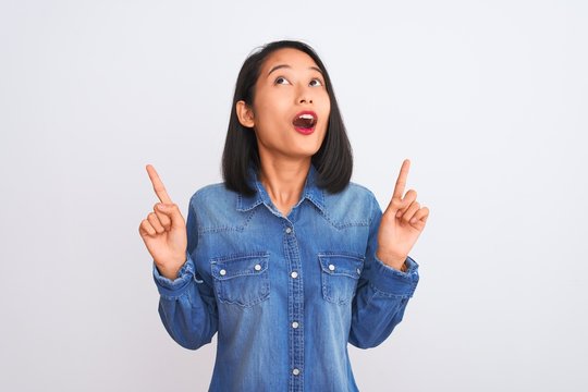Young beautiful chinese woman wearing denim shirt standing over isolated white background amazed and surprised looking up and pointing with fingers and raised arms.