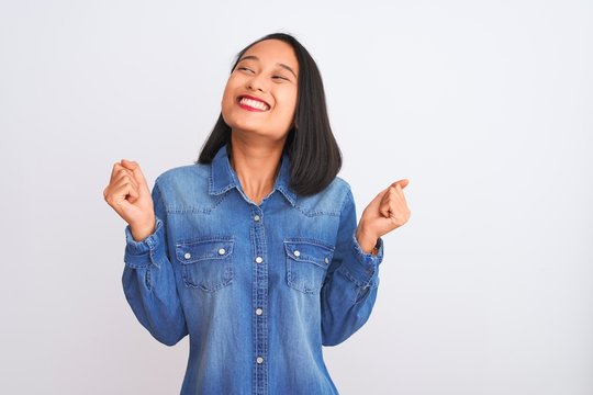 Young beautiful chinese woman wearing denim shirt standing over isolated white background celebrating surprised and amazed for success with arms raised and open eyes. Winner concept.