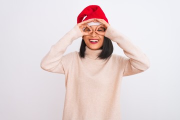 Young beautiful chinese woman wearing Christmas Santa hat over isolated white background doing ok gesture like binoculars sticking tongue out, eyes looking through fingers. Crazy expression.