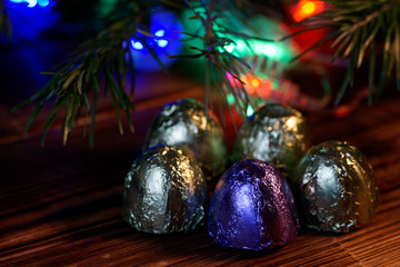 Five sweets in multi-colored foil lie on a wooden surface. Near the Christmas tree and multi-colored LED lights. Night magic. Macro. Bokeh, volume and shallow depth of field