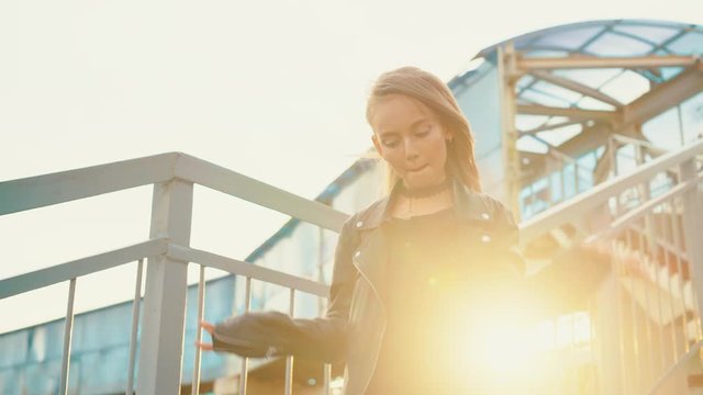 Stylish girl in leather jacket dancing on stairs outdoor. Low angle view of fashionable teenage girl with bright makeup wearting black leather jacket and dancing on steps in urban cityscape sunset.