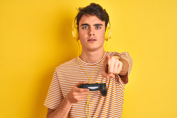 Teenager boy playing video games using gamepad over isolated yellow background pointing with finger to the camera and to you, hand sign, positive and confident gesture from the front © Krakenimages.com