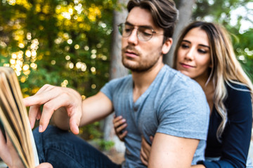 Fototapeta premium A man's hand pointing to a book, with couple embraced in the background out of focus, in a park.