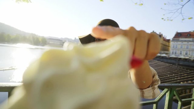 Wide angle view woman in hat taking selfie with local sweets trdelnik or Trdlo, Girl on embankment enjoying summer day, she showing buns against cityscape, laughing, pulling faces to camera 