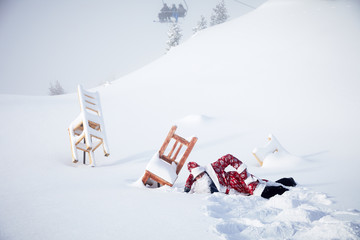 Santa Claus laying in white deep snow near wooden chairs front of skiing lift