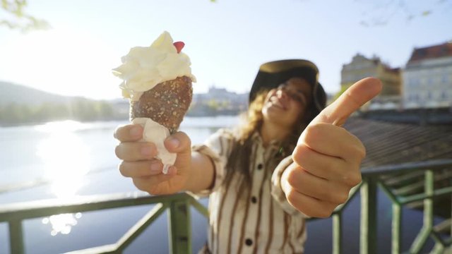 Wide angle view woman with local sweets trdelnik or Trdlo. Tourist Girl enjoying summer day, happily showing thumbs on outstretched hand and buns against cityscape, laughing, pulling faces to camera 