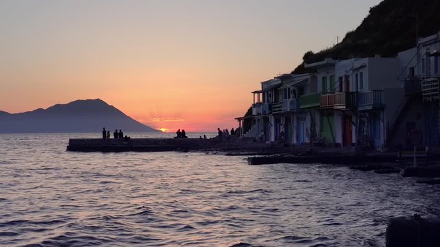 People watching colorful sunset sitting on pier in Klima fishing village on Milos island. Tourists on seaside enjoying amazing scene of sun setting behind the mountain. Romantic evening in Greece