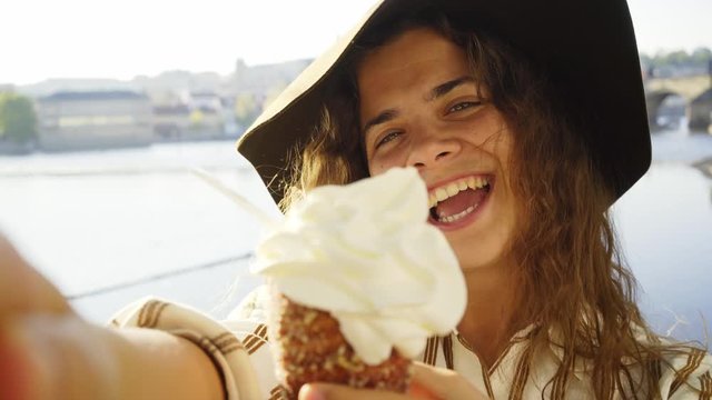 Close-up portrait woman in hat taking selfie with local sweets trdelnik or Trdlo, POV girl on embankment enjoying summer day, she showing buns against city sights, laughing, pulling faces to camera 