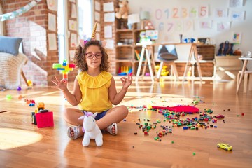 Beautiful toddler wearing glasses and unicorn diadem sitting on the floor at kindergarten relax and smiling with eyes closed doing meditation gesture with fingers. Yoga concept.