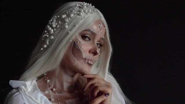 Studio portrait shot of a girl with a sweet pearl skull makeup in a room with black walls, image of a dead bride for Halloween autumn party, a woman with white long straight hair in a wedding dress