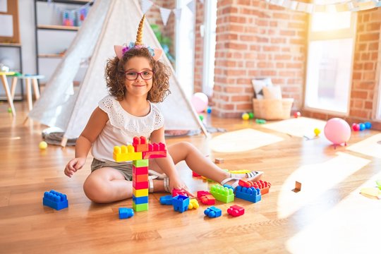 Beautiful toddler wearing glasses and unicorn diadem sitting playing with building blocks smiling at kindergarten