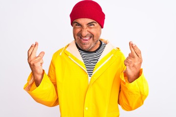 Naklejka premium Middle age man wearing rain coat and woolen hat standing over isolated white background gesturing finger crossed smiling with hope and eyes closed. Luck and superstitious concept.