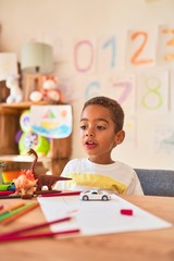 Fototapeta premium Beautiful african american toddler sitting painting car toy using marker pen on desk at kindergarten