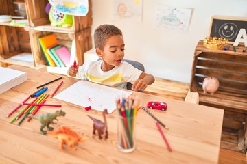 Beautiful african american toddler sitting drawing using paper and pencils on desk at kindergarten