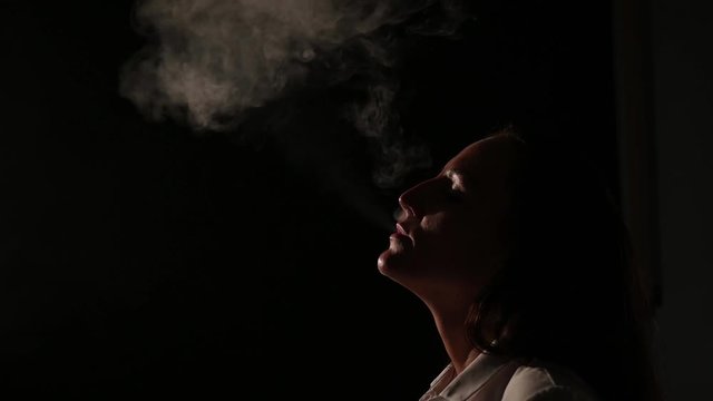 Pensive brunette in a white shirt smokes a hookah in the dark. Portrait of a woman smoking a vape, hovering. Black background. Electronic Cigarette. Bad habit.