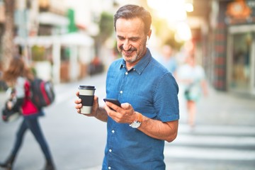 Middle age handsome businessman standing on the street using smartphone and earphones drinking coffee