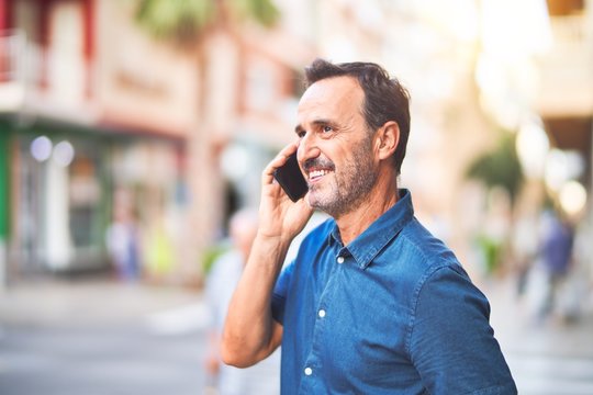 Middle age handsome businessman standing on the street talking on the smartphone smiling