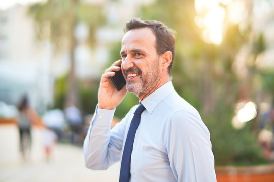 Middle age handsome businessman standing on the street talking on the smartphone smiling