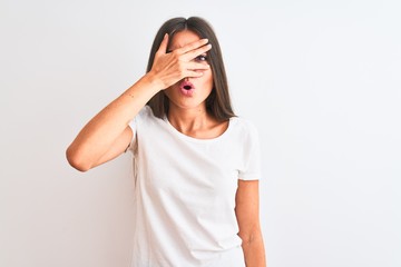 Young beautiful woman wearing casual t-shirt standing over isolated white background peeking in shock covering face and eyes with hand, looking through fingers with embarrassed expression.