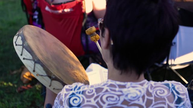 Sacred drums at spiritual singing group. A mature lady with short dark hair is seen from behind, playing a sacred native handheld drum during a gathering of people celebrating native and shaman music.