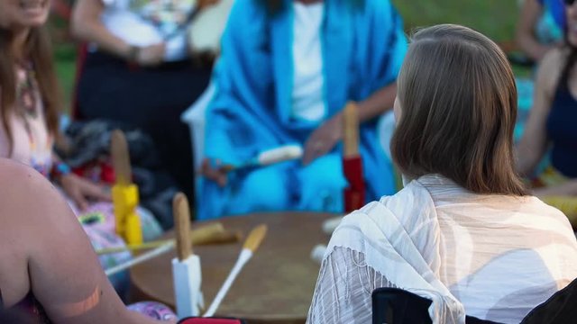 Sacred drums at spiritual singing group. Closeup and rear footage of a young woman wearing a white shawl, sat in a singing circle as people gather to celebrate shamanic and native music and culture.