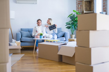 Young beautiful couple with dog sitting on the sofa holding blackboard with message at new home around cardboard boxes