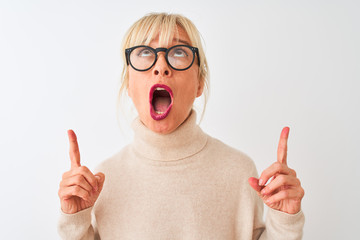 Middle age woman wearing turtleneck sweater and glasses over isolated white background amazed and surprised looking up and pointing with fingers and raised arms.