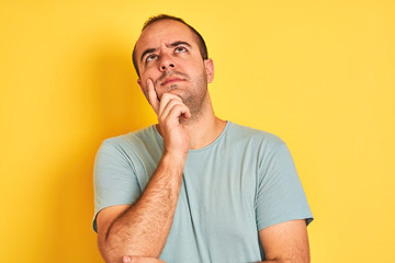 Young man wearing green casual t-shirt standing over isolated yellow background with hand on chin thinking about question, pensive expression. Smiling with thoughtful face. Doubt concept.