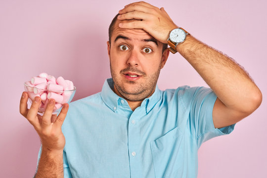 Young man holding bowl with marshmallows standing over isolated pink background stressed with hand on head, shocked with shame and surprise face, angry and frustrated. Fear and upset for mistake.