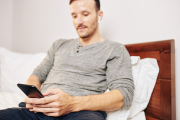 Young man sitting in bedroom, texting friends and checking social media, focus on smartphone