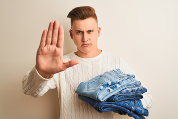 Young handsome shopkeeper man holding folded jeans over isolated white background with open hand doing stop sign with serious and confident expression, defense gesture