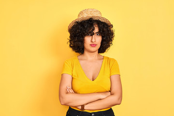 Young arab woman with curly hair wearing t-shirt and hat over isolated yellow background skeptic and nervous, disapproving expression on face with crossed arms. Negative person.