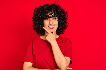 Fototapeta na wymiar Young arab woman with curly hair wearing casual t-shirt over isolated red background looking confident at the camera with smile with crossed arms and hand raised on chin. Thinking positive.