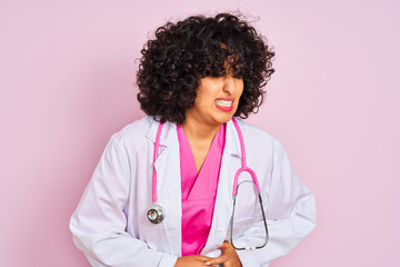 Young arab doctor woman with curly hair wearing stethoscope over isolated pink background with hand on stomach because indigestion, painful illness feeling unwell. Ache concept.