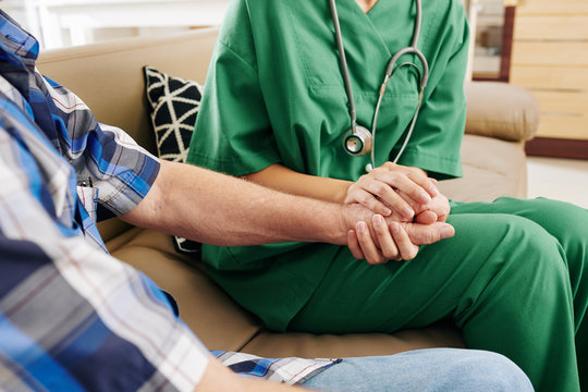 Close-up image of nurse in green scrubs holding hand of elderly patient when visiting him at home and talking about recovery process