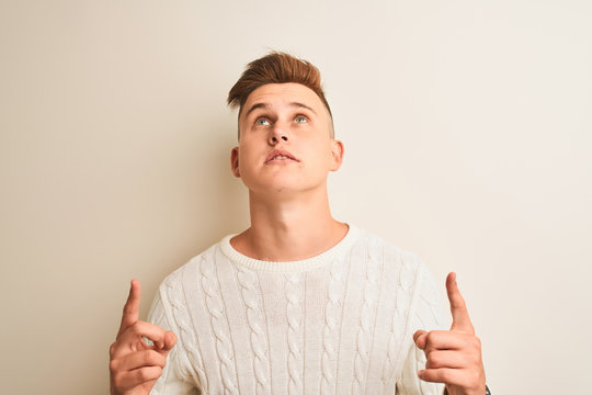 Young handsome man wearing winter sweater standing over isolated white background amazed and surprised looking up and pointing with fingers and raised arms.