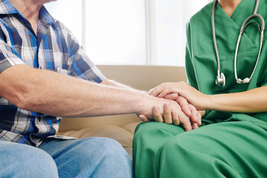 Close-up image of caregiver holding hands of elderly patient to comfort him during home visit