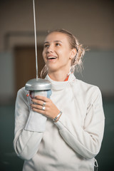 A young smiling woman fencer standing in the gym with a sword up and looking at it