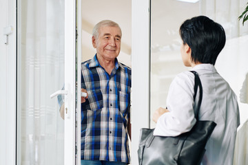 Smiling senior man in plaid shirt opening glass door to invite caregiver to house