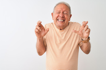 Senior grey-haired man wearing striped t-shirt standing over isolated white background gesturing finger crossed smiling with hope and eyes closed. Luck and superstitious concept.