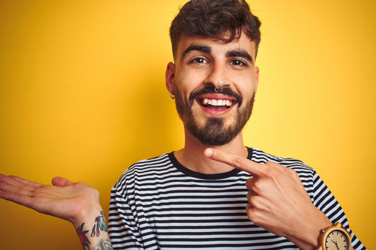 Young man with tattoo wearing striped t-shirt standing over isolated yellow background amazed and smiling to the camera while presenting with hand and pointing with finger.