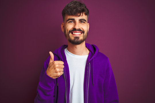 Young man with tattoo wearing striped polo standing over isolated yellow background doing happy thumbs up gesture with hand. Approving expression looking at the camera with showing success.