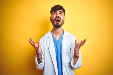 Young doctor man with tattoo standing over isolated yellow background crazy and mad shouting and yelling with aggressive expression and arms raised. Frustration concept.