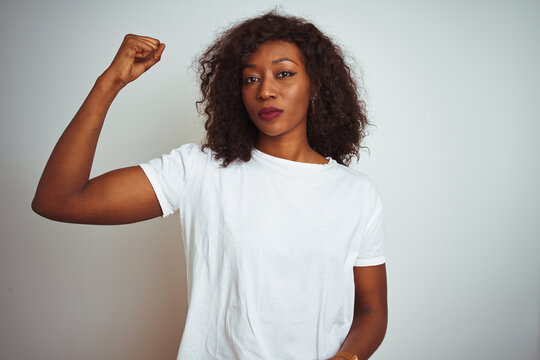 Young african american woman wearing t-shirt standing over isolated white background Strong person showing arm muscle, confident and proud of power